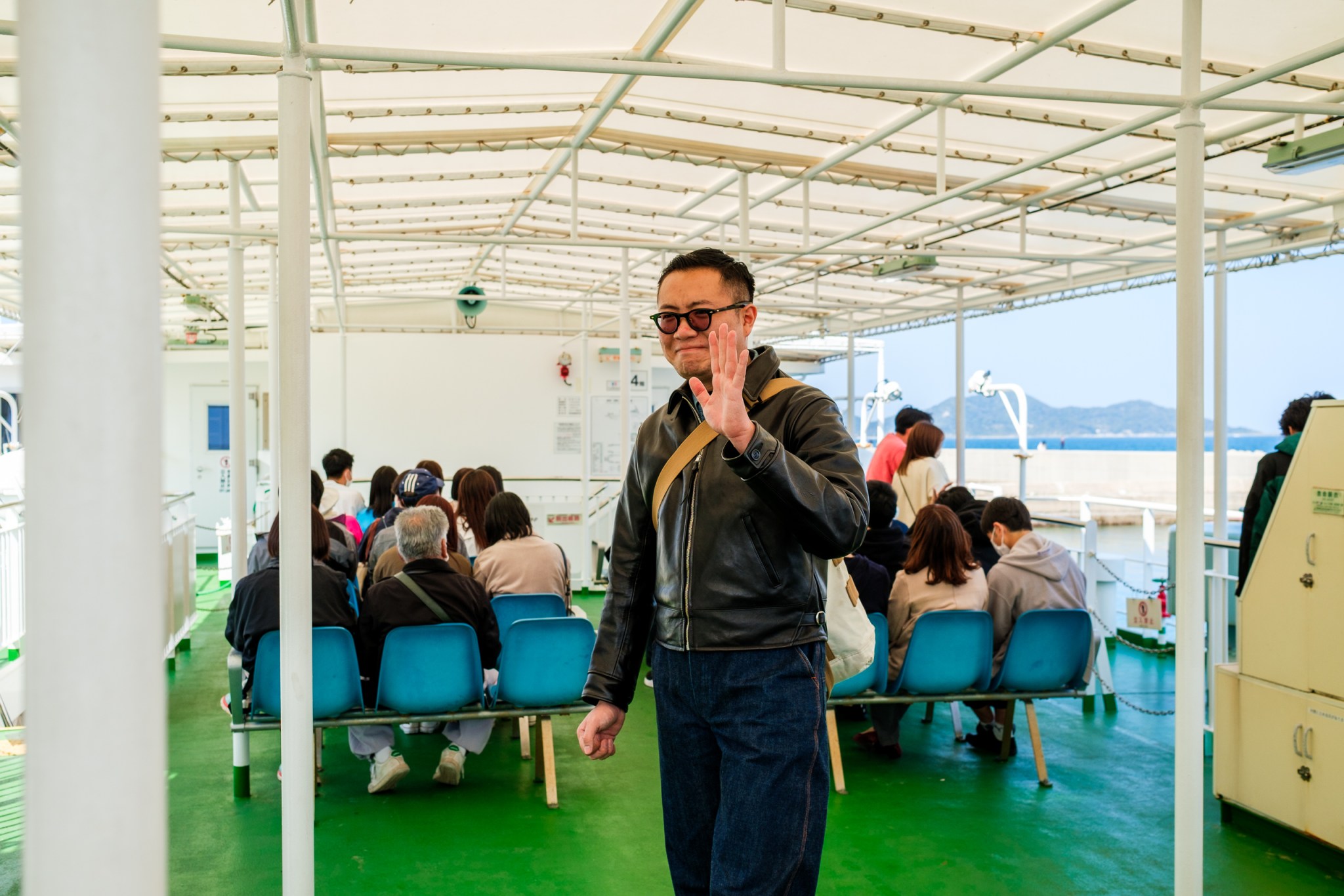 一位男子在船上与其他乘客交谈，他正在挥手致意。背景中可以看到其他乘客坐在椅子上，海洋远处隐约可见。
