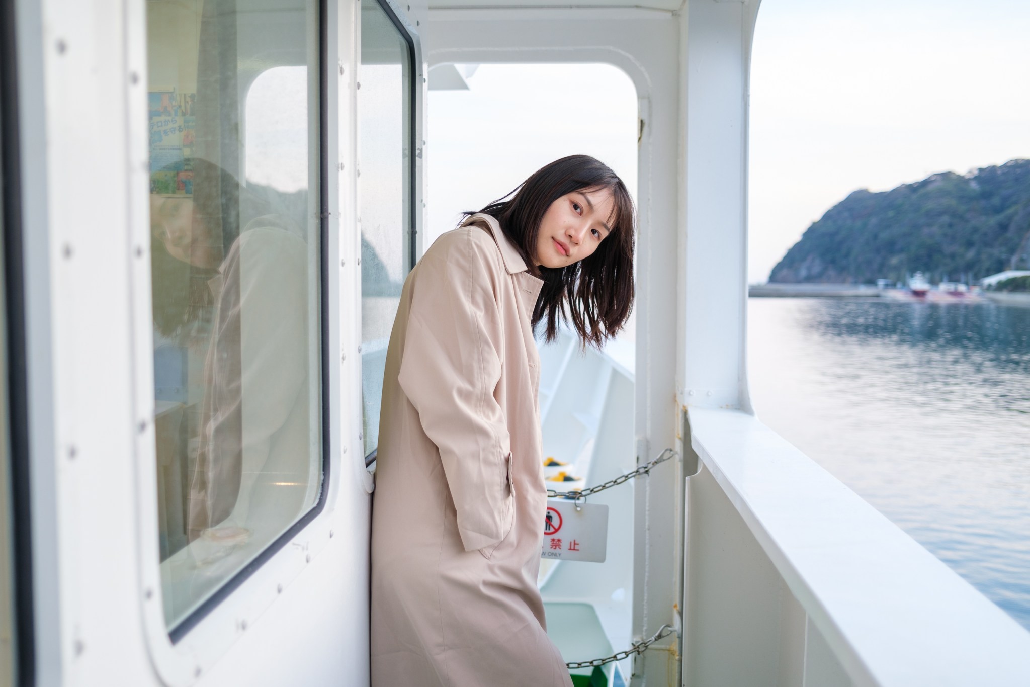 穿着米色外套的年轻女性在船舱内靠在窗边，凝视着外面的海景。