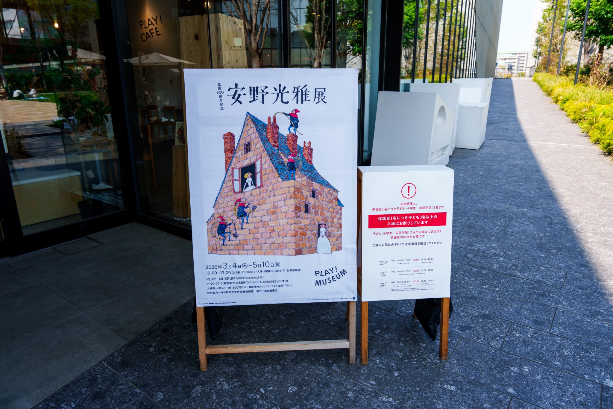 展示海报展示安野光雅的艺术作品,旁边有安全注意事项的告示牌,背景为现代建筑外观。